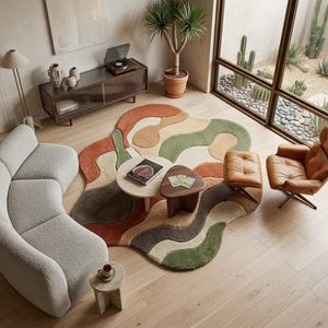 May include: An irregular, organic-shaped area rug featuring a wavy, multi-colored pattern in shades of terracotta, sage green, cream, and dark brown. The rug has a textured, tufted pile surface and is positioned on a light-colored wooden floor.