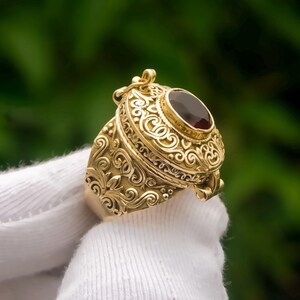 Op de afbeelding: Een gouden medaillonring met een ovaalvormige, donkerrode edelsteen in het midden. De ringband en de bovenkant van het medaillon zijn versierd met ingewikkelde, verhoogde krul- en filigraanpatronen, met een klein scharnier aan de zijkant.