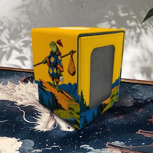 Puede incluir: Una caja para barajas amarilla con el gráfico de una rana verde que viste una túnica azul con flores amarillas, cargando un palo con un saco al hombro. La caja tiene un recorte rectangular en el lateral y una ilustración de montañas.
