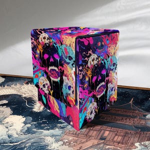 Op de afbeelding: Een vierkante deck box met een levendige, veelkleurige grafische print van schedels met druipende verfeffecten in neonroze, elektrisch blauw en dieppaars tegen een donkere achtergrond, met een veilig deksel en een gestructureerde rechthoekige vorm.