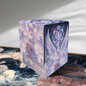 May include: A cube-shaped card storage box with a fantasy angel illustration. The box features a holographic design with a seated angel with large wings on the front and side panels. The box is black on the top and has a magnetic closure.