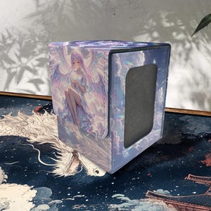 May include: A cube-shaped card storage box with a fantasy angel illustration. The box features a holographic finish with a flap opening. The artwork depicts a seated angel with large wings, in shades of purple and blue.