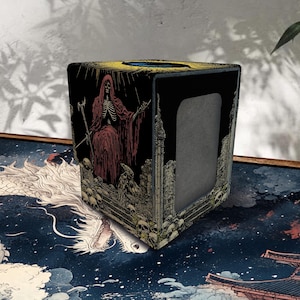 Op de afbeelding: Een vierkante deckbox met een donkere illustratie van een skeletfiguur met een kap en rode mantel die een staf vasthoudt, omringd door schedels en stenen ruïnes. De doos heeft een zwarte achtergrond, een gele bovenrand en een rechthoekige uitsparing.