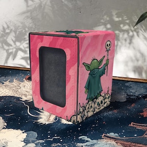 May include: Pink cube-shaped card storage box with a cutout window. The box features a cartoon illustration of a green alien-like character in a robe holding a staff with a skull on top. Skulls are at the base of the character.