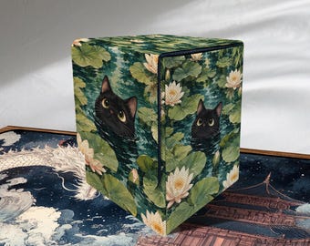 May include: A square box featuring a printed pattern of two black cats peeking out from a pond filled with green lily pads and white water lily flowers. The box has sharp edges and a smooth surface, displaying a detailed botanical and animal scene.