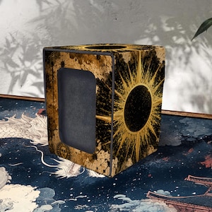 May include: A decorative box with a sunburst design in gold and black. The box has a rectangular opening on one side and is placed on a patterned surface. The box's design features a sun with radiating lines.
