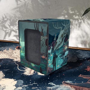 May include: A teal, square storage box with a fantasy landscape design featuring a deer skull, figures, and a forest scene. The box has a rectangular opening and a dark gray interior.