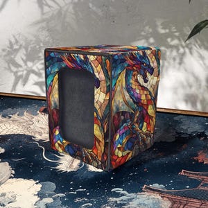 May include: A square, stained-glass-style box featuring a colorful dragon design. The dragon is depicted with wings and is rendered in shades of blue, red, orange, and yellow. The box has a dark interior and a rectangular opening.