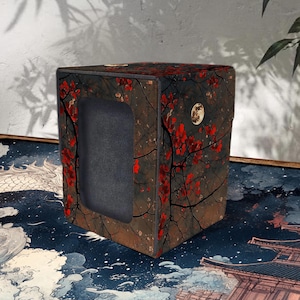 Puede incluir: Una caja de almacenamiento cuadrada con un fondo oscuro y un diseño de inspiración japonesa. La caja presenta un patrón de flores y ramas rojas. Un acento dorado circular es visible en el lateral. La caja tiene una abertura rectangular en la parte delantera.