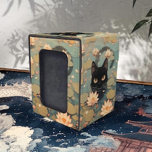 May include: A square storage box with a black cat design. The box features a teal background with a pattern of lily pads and flowers. A black cat with yellow eyes is featured on the side. The box has a rectangular opening on one side.