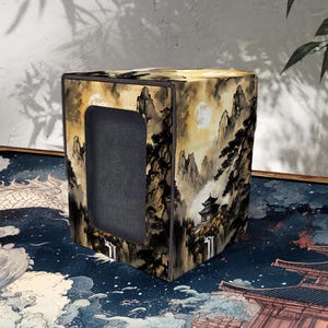 May include: A square storage box with a landscape design featuring mountains, trees, and a full moon. The box has a dark grey interior and a rectangular opening on one side. The colour palette includes shades of brown, beige, and black.