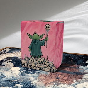 Peut inclure: Boîte de rangement pour cartes rose avec un personnage de dessin animé vert tenant un bâton, debout sur des crânes. Le personnage porte une robe à capuche vert foncé. La boîte a une texture.