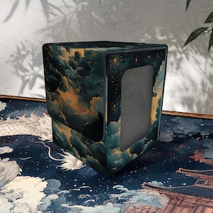 Puede incluir: Una caja de almacenamiento de tarjetas cuadrada con un diseño celestial. La caja presenta un paisaje de nubes verde oscuro y dorado con un cielo estrellado en un lado. La caja tiene una abertura rectangular en la parte delantera.