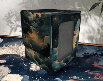 Puede incluir: Una caja de almacenamiento de tarjetas cuadrada con un diseño celestial. La caja presenta un paisaje de nubes verde oscuro y dorado con un cielo estrellado en un lado. La caja tiene una abertura rectangular en la parte delantera.