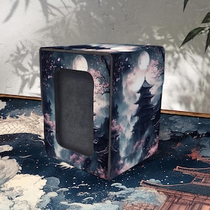 May include: A square storage box with a Japanese-inspired design. The box features a dark blue and pink landscape with a pagoda, cherry blossoms, and a full moon. The box has a rectangular opening on one side.