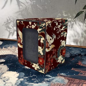 May include: A burgundy box with a rectangular opening, adorned with a Japanese-inspired design. The pattern features white phoenixes, dragons, and floral elements. The box sits on a blue and white patterned surface.