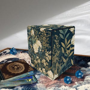 May include: A floral-patterned card storage box with a dark blue background and cream-colored flowers. The box is surrounded by blue dice and a deck of cards. The box is designed to hold trading cards.