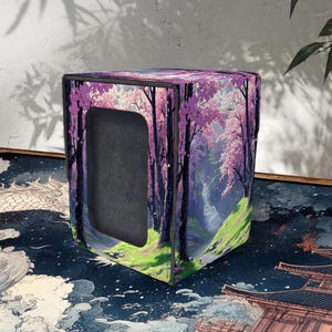May include: A cube-shaped storage box with a Japanese landscape design. The box has a black interior and a vibrant exterior featuring cherry blossom trees, a waterfall, and a green grassy area. The colour palette includes shades of purple, pink, and green.