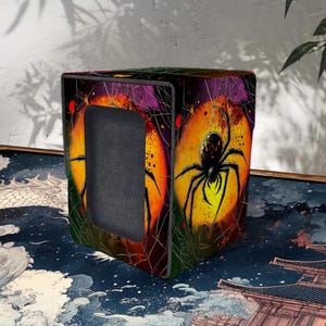 Puede incluir: Una caja cuadrada negra y naranja con una gran ilustración de una araña negra. La araña está sobre un fondo amarillo y naranja, con un diseño de telaraña. La caja tiene una abertura rectangular.