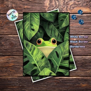 Op de afbeelding: Een digitale kunstprint met een groene kikker die door weelderige groene bladeren gluurt. De kikker heeft grote, oranje ogen. De print is verkrijgbaar met een witte rand, zwarte rand of zonder rand. De woorden "Matte & Foil" zijn ook zichtbaar.
