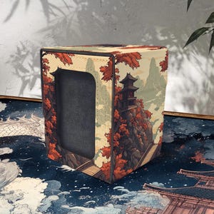 May include: A cube-shaped storage box with a Japanese-inspired landscape design. The box features a detailed illustration of a pagoda on a cliff surrounded by autumn foliage. The color palette includes shades of red, orange, beige, and green.