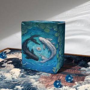 May include: A blue card storage box featuring a koi fish design. The artwork depicts a black and white koi fish swimming in a pond with lily pads. The box is surrounded by blue dice and a decorative mat with a wave pattern.