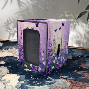 Peut inclure: Un étui à boîte de mouchoirs carré avec un motif floral dans les tons de violet, rose et blanc. Le couvercle présente une silhouette de chat noir et des papillons. La boîte a une ouverture rectangulaire.