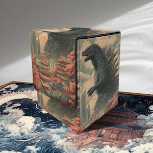 Puede incluir: Caja de almacenamiento cúbica con una ilustración detallada de Godzilla en un paisaje de inspiración japonesa. La caja tiene un borde azul y un cierre de solapa. La obra de arte incluye pagodas, flores de cerezo y un paisaje urbano.