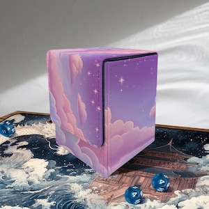 Könnte beinhalten: Eine quadratische Würfelbox mit einem Himmelsdesign. Die Box zeigt einen violetten Himmel, flauschige Wolken und Sterne. Sie hat einen Klappverschluss und einen schwarzen Rand. Drei blaue Würfel liegen verstreut.