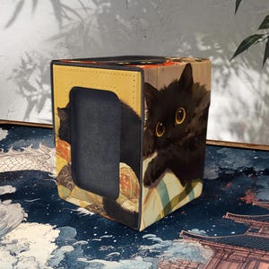 Peut inclure: Boîte de rangement pour cartes ornée d'une illustration de chat noir. La boîte a un cadre jaune et noir avec une découpe. Le chat a de grands yeux jaunes et se détache sur un fond d'art japonais traditionnel.