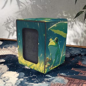 May include: A square, teal card storage box with a large rectangular opening. The box features a cartoon illustration of a green frog under a leaf in a rain shower. The box has a black interior and a yellow and green gradient on the bottom.