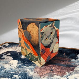 May include: A cube-shaped storage box featuring a Japanese-inspired design. One side displays a gray cat with yellow eyes, wrapped in a red and white blanket. Other sides show a ball of yarn and a traditional Japanese scene with waves and a pagoda.