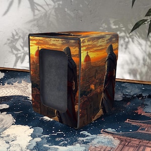 May include: A decorative box with a sunset cityscape design, featuring figures in hooded cloaks. The box has a rectangular opening. The artwork shows a detailed architectural scene with a warm colour palette.
