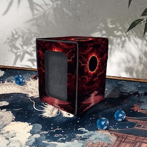 Könnte beinhalten: Ein würfelförmiger Würfelbehälter mit einem feurigen roten und schwarzen Design, das ein zentrales Sonnenfinsternisbild zeigt. Die Box hat eine rechteckige Öffnung und ist von einem dunklen, wolkigen Himmelmuster umgeben. Mehrere blaue Würfel liegen um die Box herum.