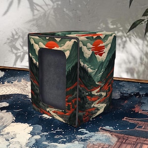 May include: A square storage box with a landscape design featuring mountains, a red sun, and a river. The box has a rectangular opening and is primarily green, red, and white. The box is sitting on a blue surface with a Japanese-style wave pattern.