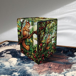 Puede incluir: Una caja de almacenamiento cúbica con un diseño de vidriera que representa ardillas en un árbol con bayas rojas. La caja tiene un borde azul oscuro y está colocada sobre una superficie decorativa con un diseño de olas y edificios de estilo japonés.