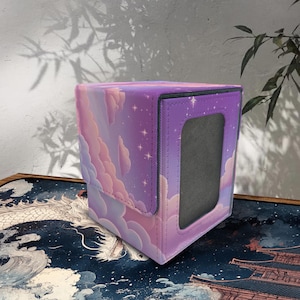 May include: A square, purple card storage box with a flap closure. The box features a dreamy design of pink and white clouds, stars, and a purple sky. The box has a black trim and a rectangular window.