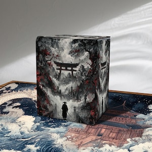 May include: A cube-shaped storage box featuring a Japanese-inspired landscape design. The artwork depicts a samurai figure, a torii gate, and a temple, with a color palette of gray, red, and black. The box is likely used for storing items.