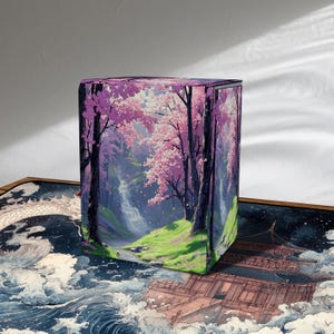 May include: A rectangular box featuring a vibrant illustration of a cherry blossom forest. The artwork depicts pink and white flowering trees, a waterfall, and a path, set against a backdrop of purple and green hues. The box is placed on a surface with a dragon and wave design.