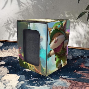 Puede incluir: Caja de almacenamiento cúbica con una ilustración de un personaje de dibujos animados en verde y marrón. La caja tiene una abertura rectangular y está sobre una superficie estampada. El diseño es vibrante y llamativo.