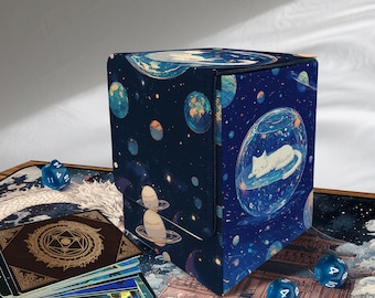 Puede incluir: Una caja de almacenamiento de cartas azul oscuro con un diseño espacial que presenta planetas, estrellas y un gato durmiente en una burbuja de vidrio. Dados azules y una baraja de cartas.