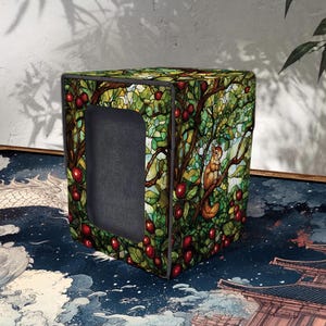 Op de afbeelding: Een kubusvormige opbergdoos met een glas-in-loodstijl ontwerp met een eekhoorn in een boom met rode appels. De doos heeft een donkergrijze binnenkant zichtbaar door een rechthoekige opening. De achtergrond bevat een blauw en wit patroon.