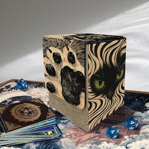 Peut inclure: Boîte de rangement de cartes en forme de cube avec un motif de visage de chat noir et d'empreinte de patte. La boîte présente un visage de chat noir avec des yeux verts d'un côté et une empreinte de patte de l'autre. Des dés bleus et un jeu de cartes entourent la boîte.