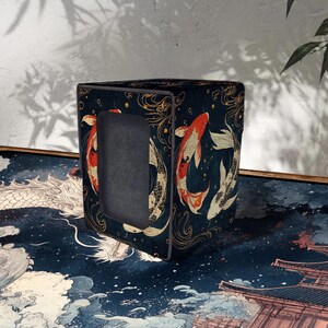 May include: A dark blue tissue box cover featuring a koi fish design in red, white, and gold. The box has a rectangular opening and is decorated with an Asian-inspired pattern. The tissue box cover is a decorative item.
