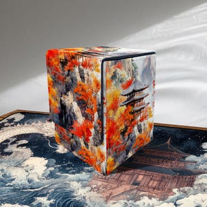 Puede incluir: Una caja de almacenamiento cuadrada con un colorido diseño de paisaje japonés. La caja presenta una escena de montaña con follaje otoñal y una pagoda. La caja está sobre una alfombra con un diseño de dragón y olas.