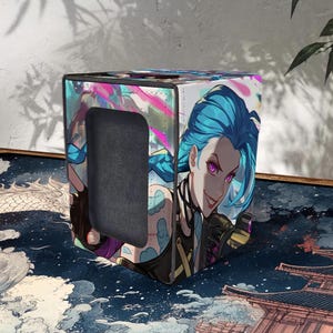Può includere: Scatola portaoggetti a forma di cubo con una vivace illustrazione di un personaggio con capelli blu e occhi viola. La scatola ha un ritaglio su un lato ed è decorata con un design colorato in stile anime. La scatola è posizionata su una superficie a motivi.