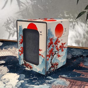 Puede incluir: Una funda de caja de pañuelos cuadrada y blanca con un diseño de paisaje japonés. La obra de arte incluye un sol rojo, montañas, árboles con hojas rojas y pájaros volando. La caja tiene una abertura gris oscura.