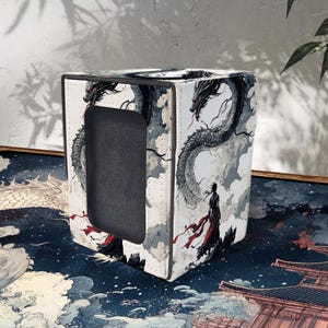 Puede incluir: Un tarjetero cúbico blanco con una ilustración detallada de un dragón y una figura con túnicas rojas contra un cielo nublado. El diseño recuerda al arte asiático tradicional. El soporte tiene una abertura negra.