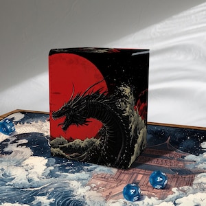 Puede incluir: Caja de almacenamiento cúbica con una ilustración de estilo japonés de un dragón negro, un sol rojo y olas. Varios dados azules están dispersos alrededor de la caja.