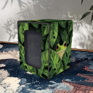 Puede incluir: Caja de almacenamiento cúbica con un patrón de hojas verdes y un diseño de rana. La caja tiene una abertura rectangular y está colocada sobre una superficie estampada. La caja mide unos 30 cm de alto.